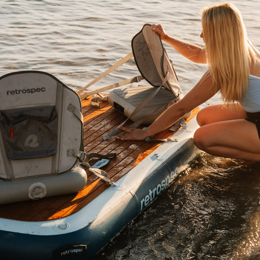 A person adjusts the seat on a inflatable dock at the water's edge with the brand name "retrospec" visible.