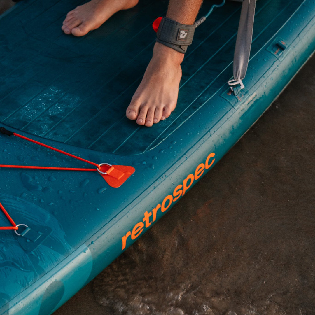 Close-up of feet on a teal Retrospec inflatable paddle board showing deck traction pad and ankle leash in shallow water.