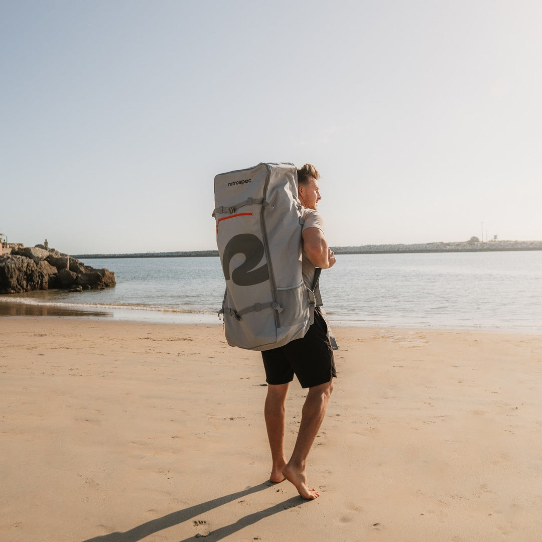 Man with retrospec paddle board bag backpack walking on beach toward water
