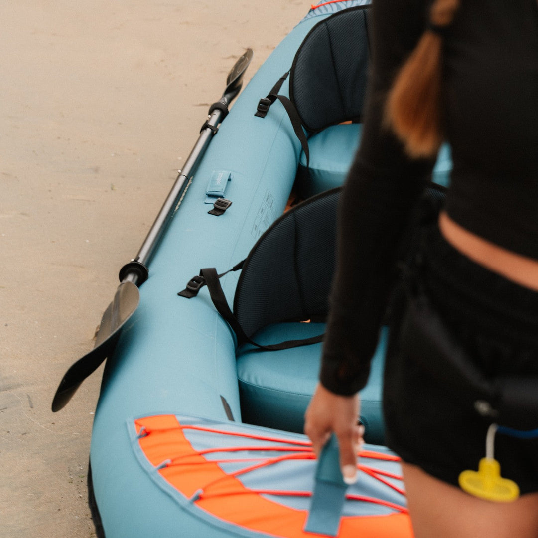 A person carries the a tandem kayak by the front handle while walking on a sandy beach.