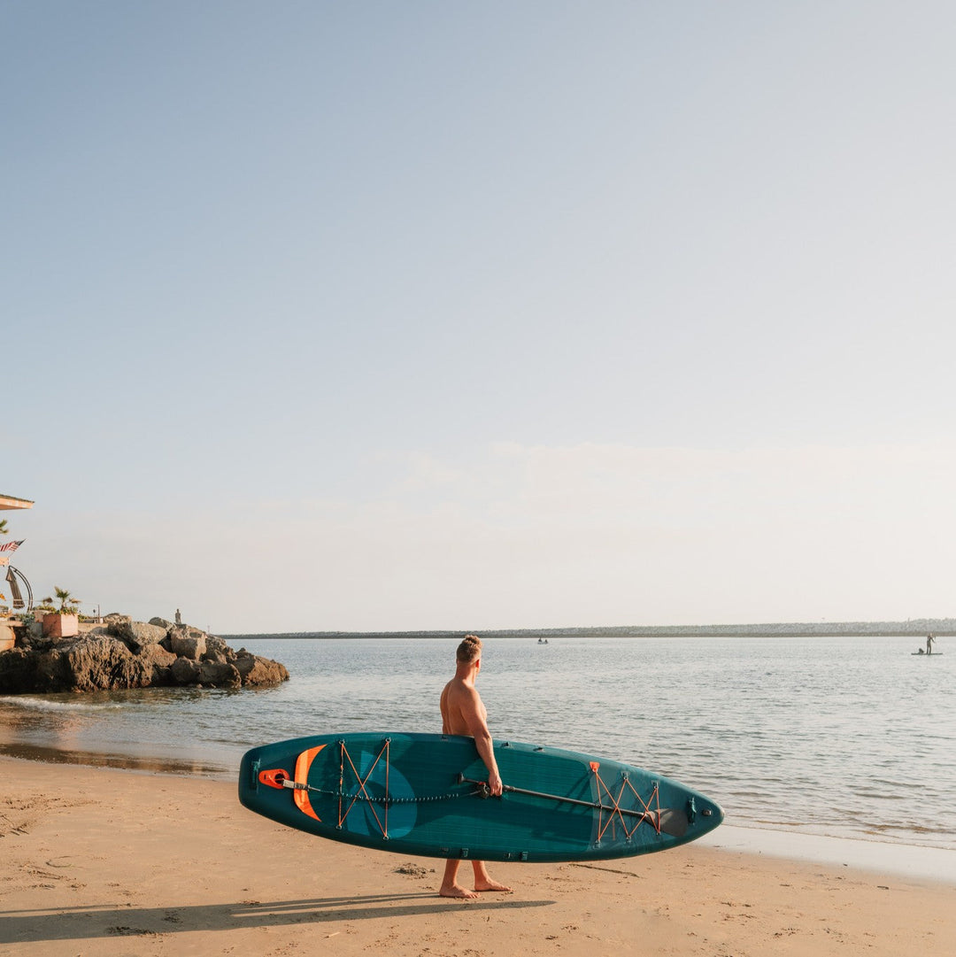 Man carrying teal retrospec Weekender Tour inflatable paddleboard on sandy beach with ocean water in background