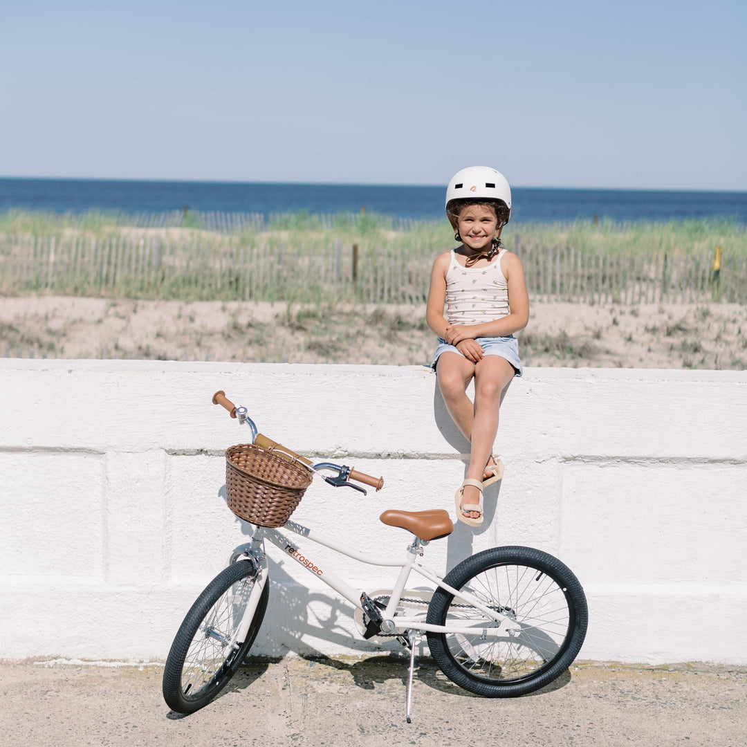 A child wearing a white helmet sits on a white wall with sand dunes and the ocean behind them.