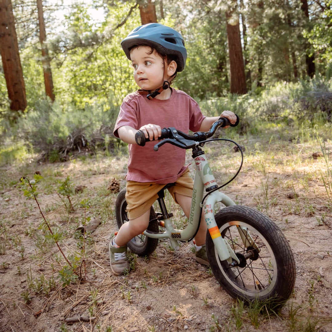 A child in a helmet rides a balance bike on a sandy trail surrounded by tall trees and greenery, enjoying the outdoors.