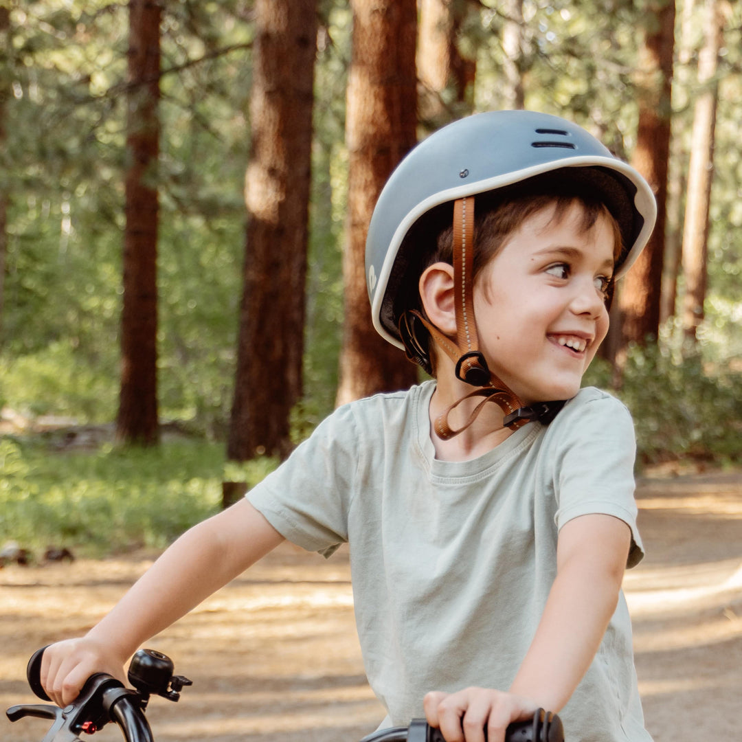A child in a light green shirt and a grey helmet rides a bike on a forest path surrounded by tall trees.