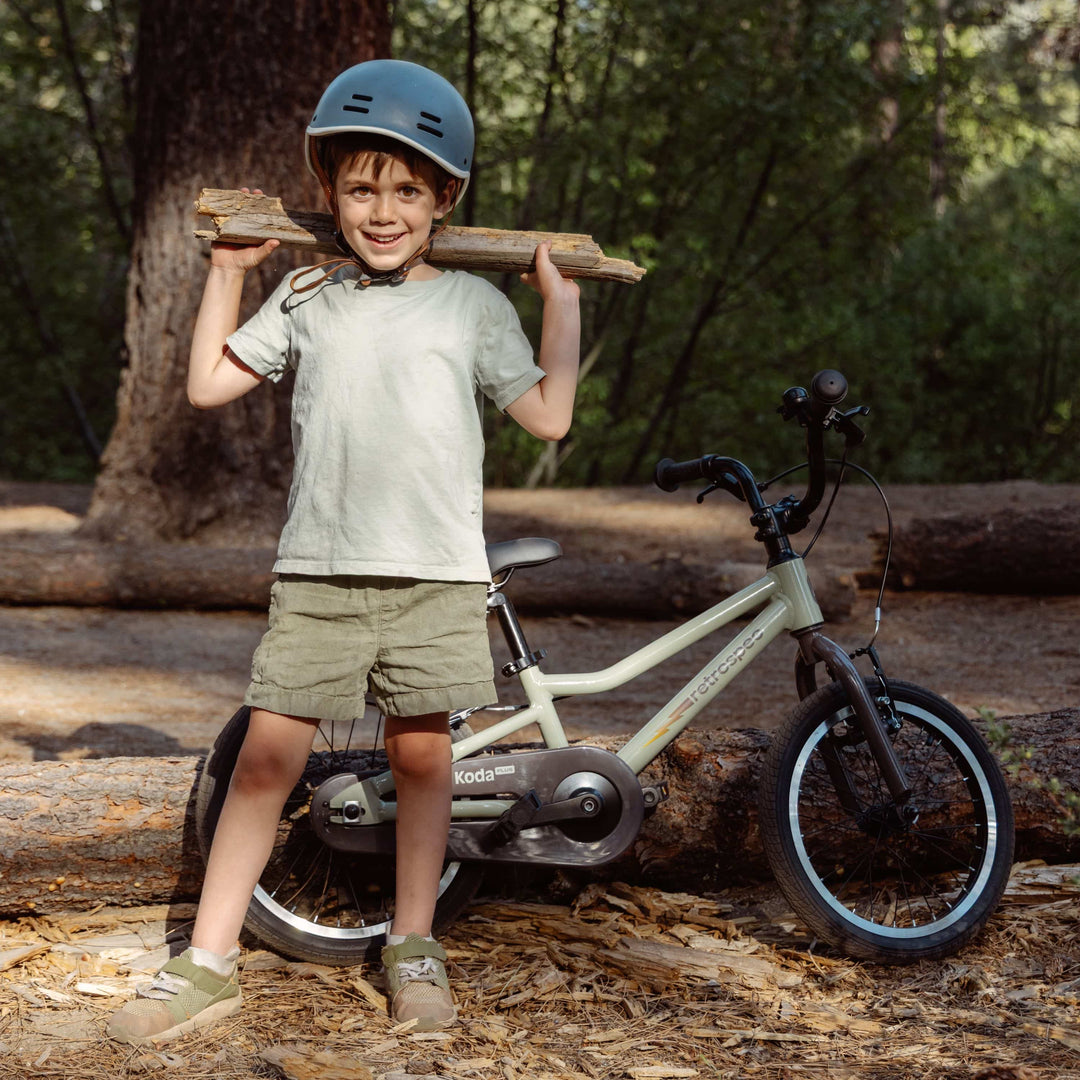 Smiling child in a helmet standing next to a sage green Retrospec Koda Plus 16 inch kids bike among fallen logs in a forest.