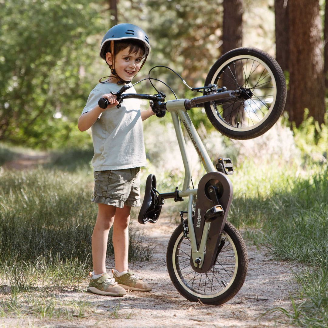 A child in a helmet lifts a bicycle on a forest path, surrounded by greenery and sunlight filtering through the trees.