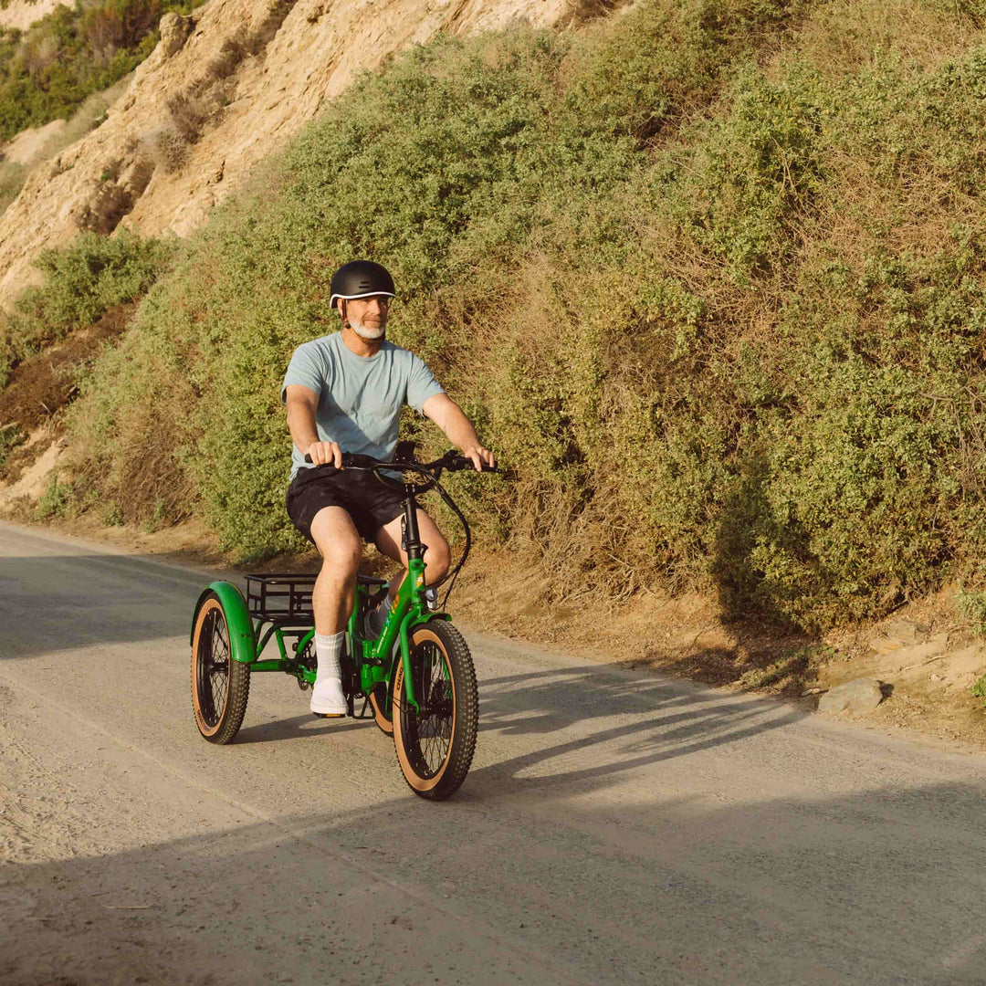 A person rides a green three-wheeled bike on a gravel path surrounded by lush greenery and rocky hills.