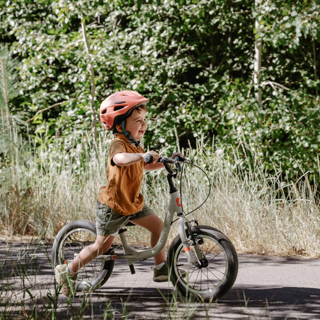 Child riding gray retrospec Cub Plus XL toddler push bike with red helmet outdoors, surrounded by green vegetation.
