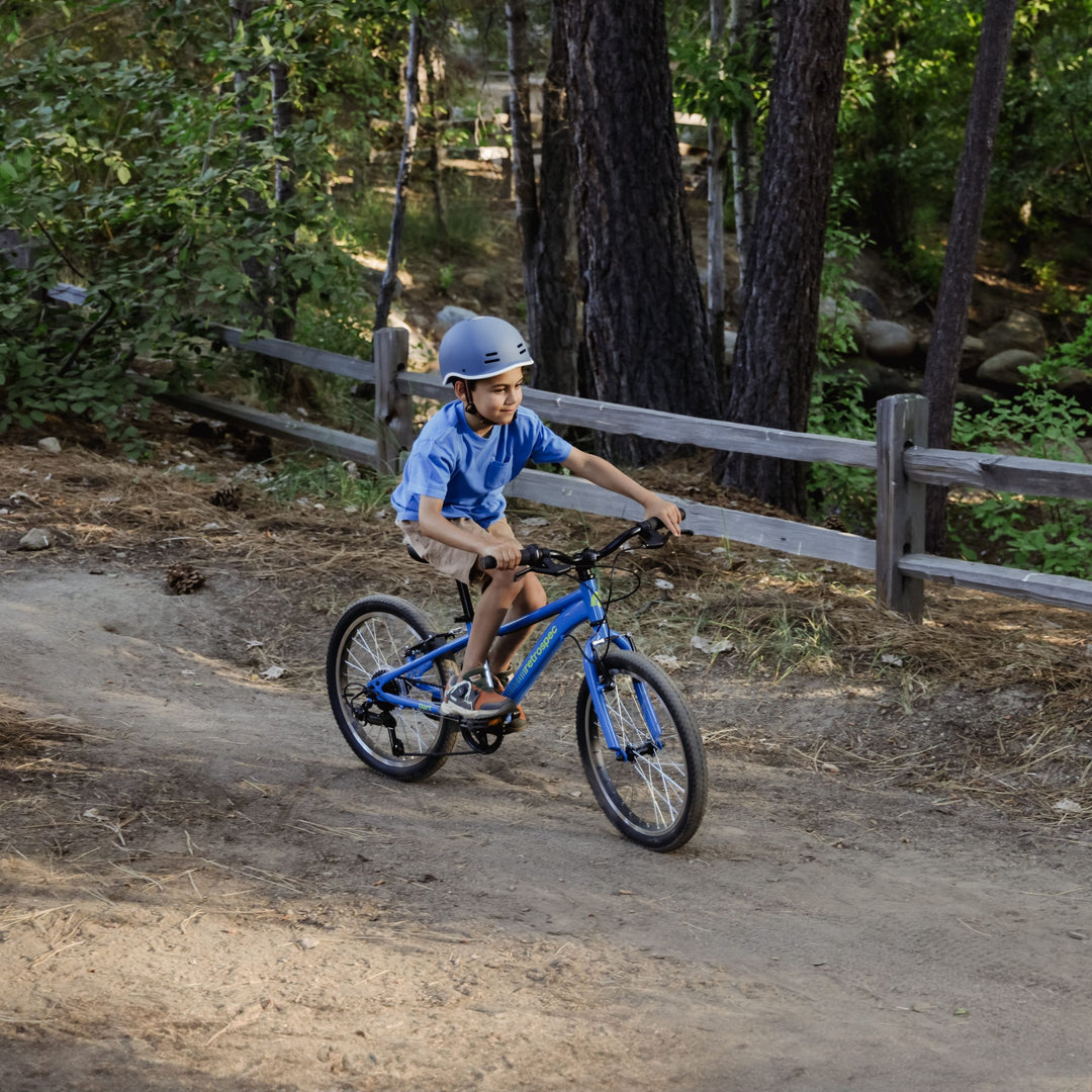 A child rides a blue bicycle on a dirt path, surrounded by green trees and a wooden fence, enjoying a sunny day outdoors.