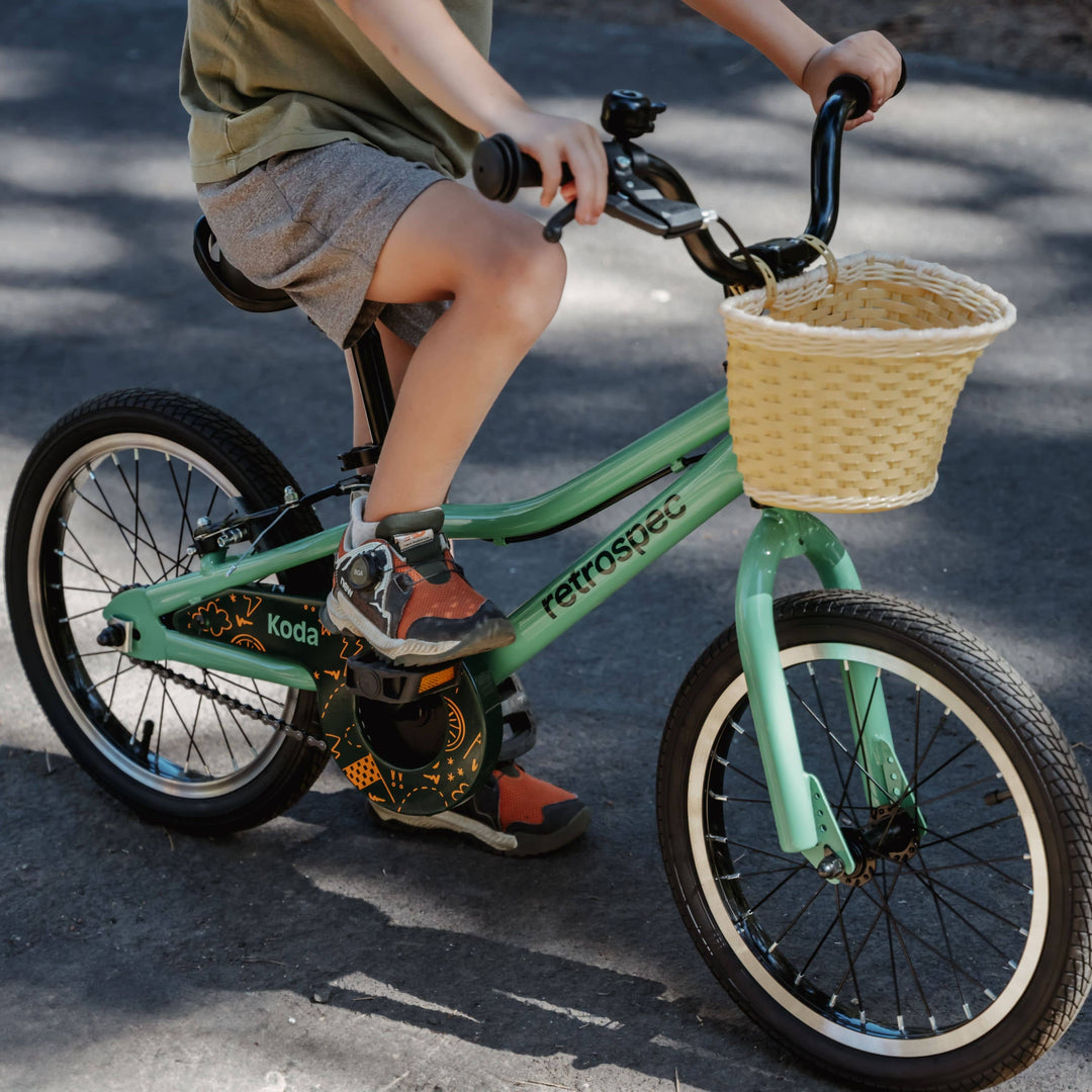 A child rides a green bike with a woven basket, wearing shorts and sporty shoes, on a sunlit road.