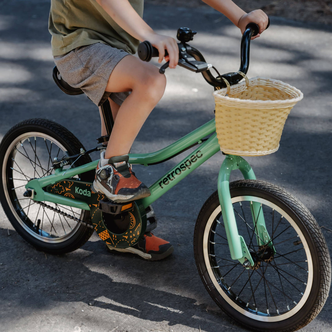 Close-up of a child's feet pedaling a green Retrospec Koda 16 inch kids bike with a wicker handlebar basket.
