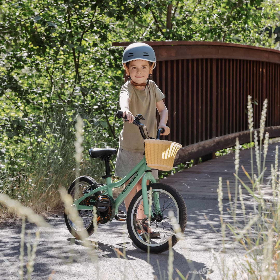 Smiling child on a green retrospec Koda bike for kids on a paved path with a wooden bridge and lush greenery in the background.