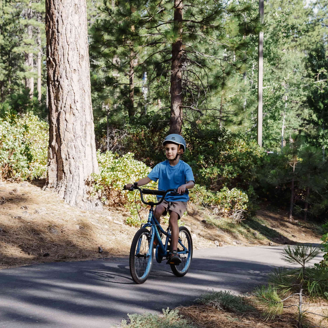 Child in a helmet riding a blue Retrospec Koda 20 inch kids bike toward the camera on a shaded forest path.