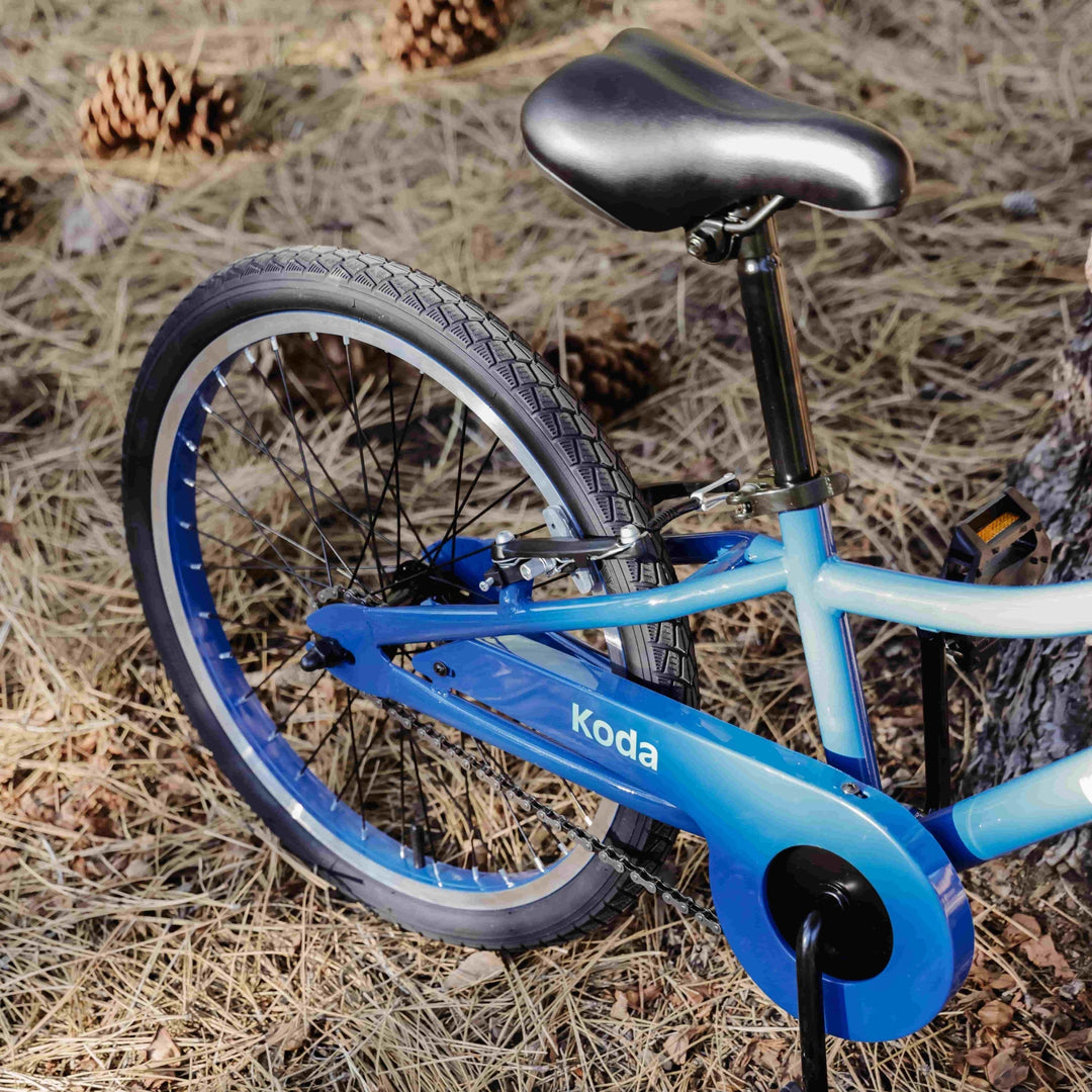 Close-up of the blue Retrospec Koda kids bike rear frame, seat, and chain showing the Koda logo among pine cones.