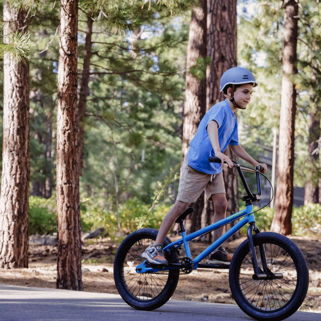 A child rides a blue BMX bike on a path through a forest, surrounded by tall trees and greenery, wearing a helmet and casual clothes.