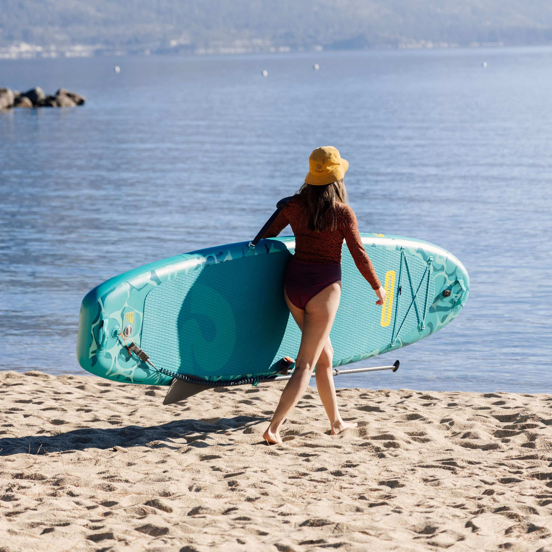 A woman in a yellow bucket hat and rust long-sleeve walks barefoot along a sandy lakeshore, carrying a teal Retrospec Paddle Board under one arm and a paddle in the other hand, with pine trees, rocky jetties, and a calm mountain lake in the background.