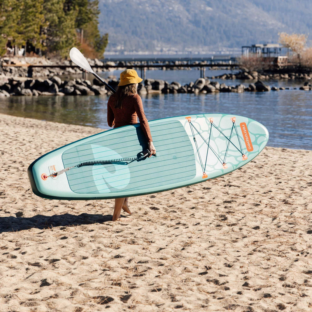 Woman carrying retrospec Weekender 10 paddle board and paddle on a sandy beach with mountain lake views.