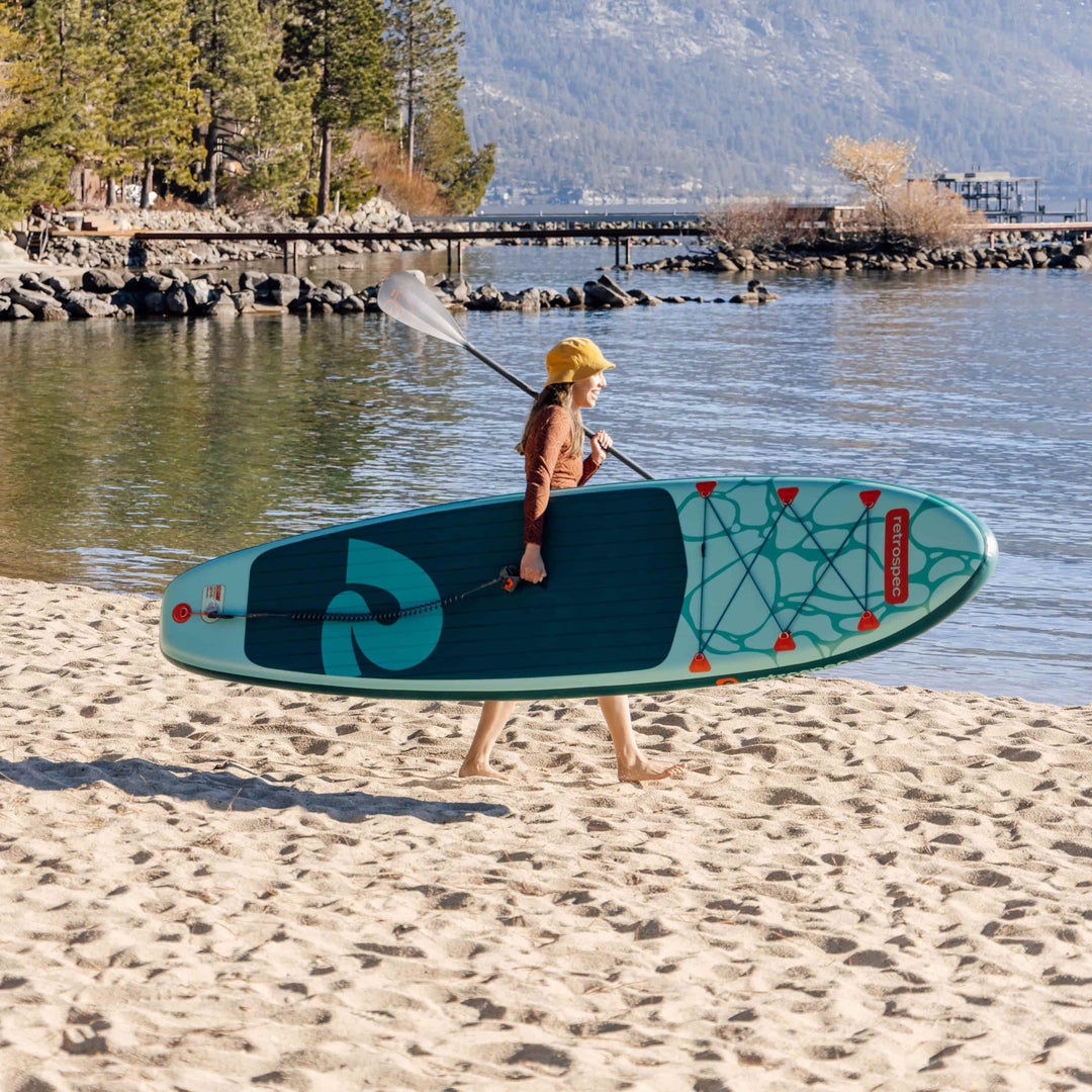 Woman in a yellow cap carrying a teal retrospec inflatable paddle board on a sandy beach overlooking a mountain lake.