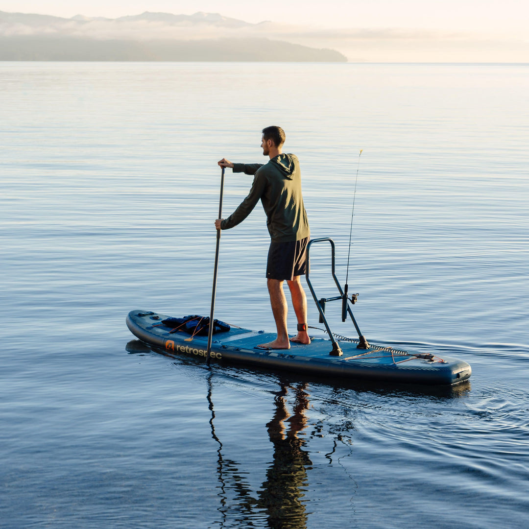 Man on retrospec June inflatable fishing kayak with paddle and fishing rod, calm water and mountain backdrop.