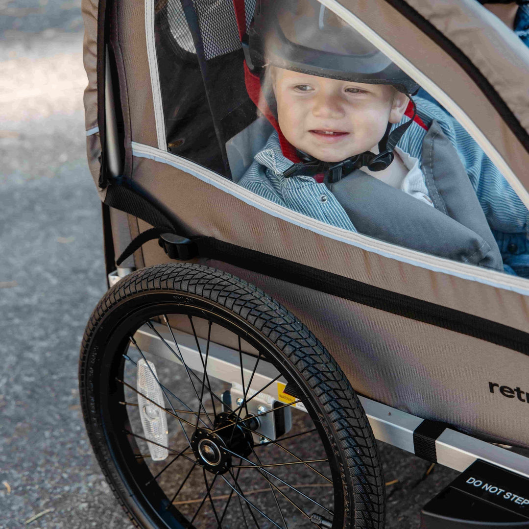 A child seated in a gray bike trailer, with a large wheel in the foreground, and sunlight illuminating the scene.