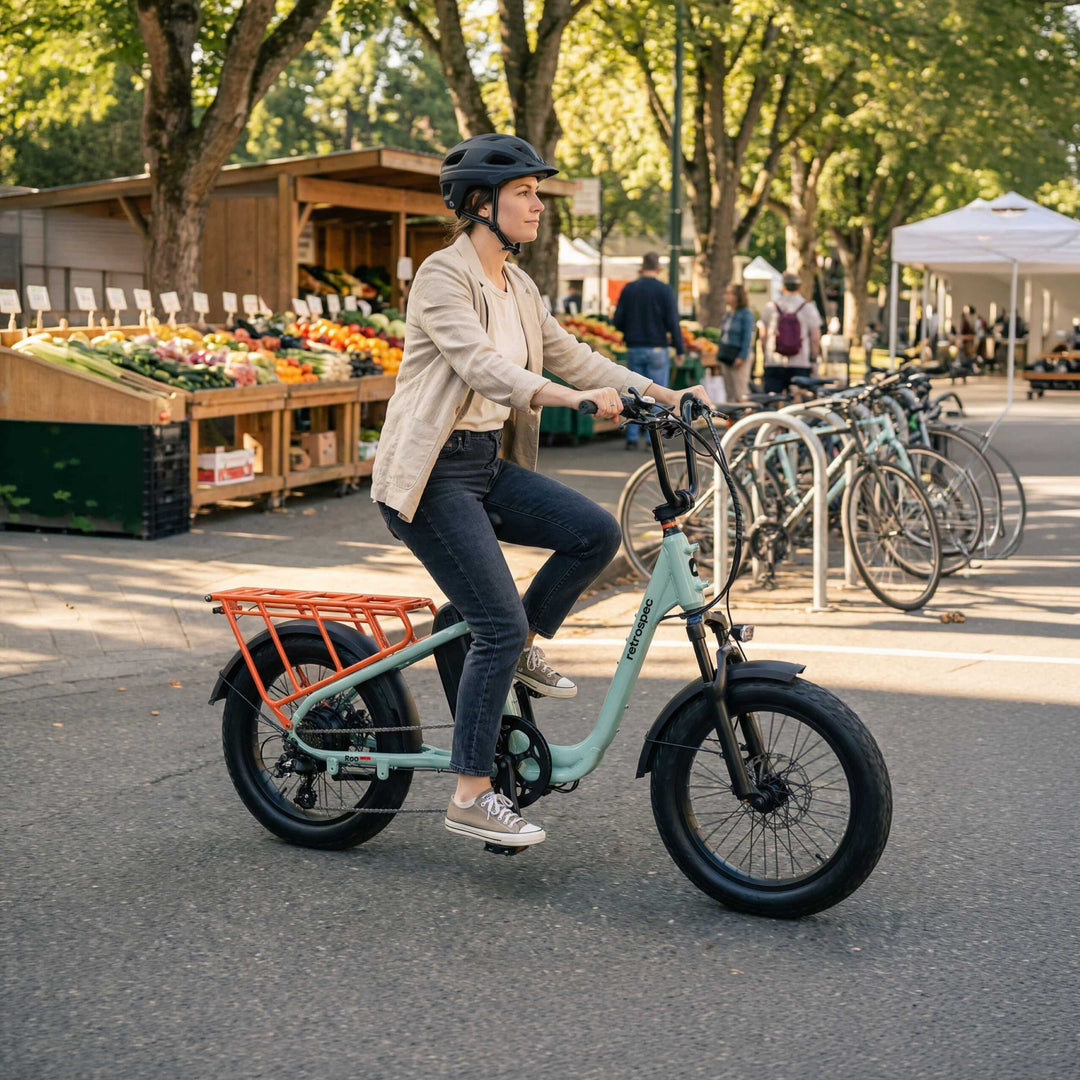 A woman wearing a helmet rides the retrospec Roo Rev XL Cargo Electric Bike in Mint green past a vibrant outdoor farmers market, with fresh produce stands, tree-lined paths, and parked bikes visible in the background.