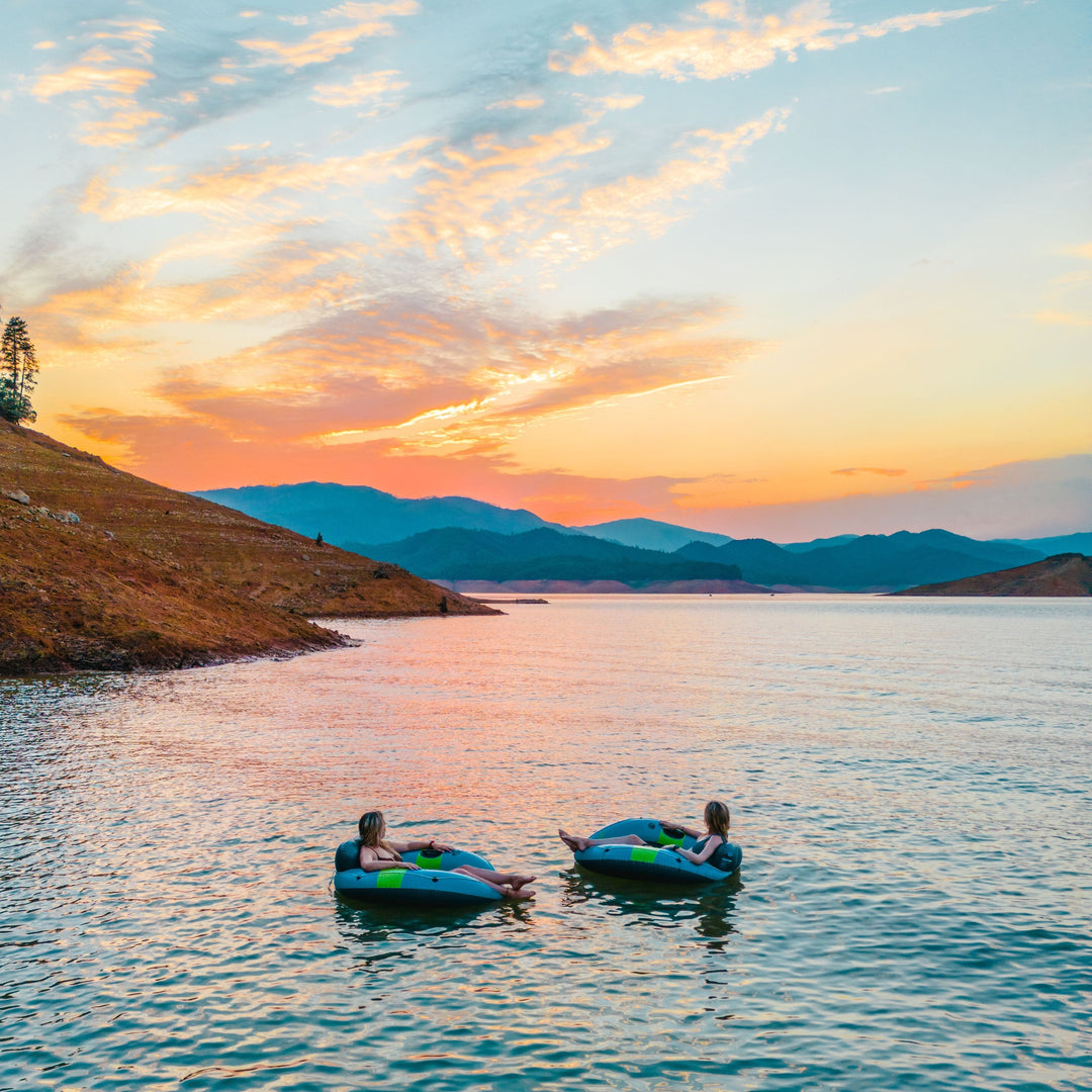 Two people relax on inflatable tubes in a calm lake at sunset, surrounded by hills and colorful sky.