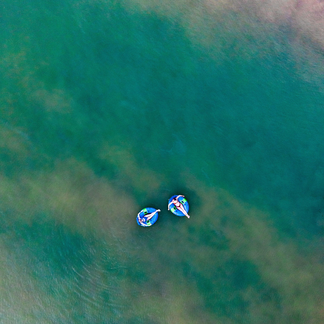 Two people relax on colorful inflatable tubes in calm, clear water, viewed from above.