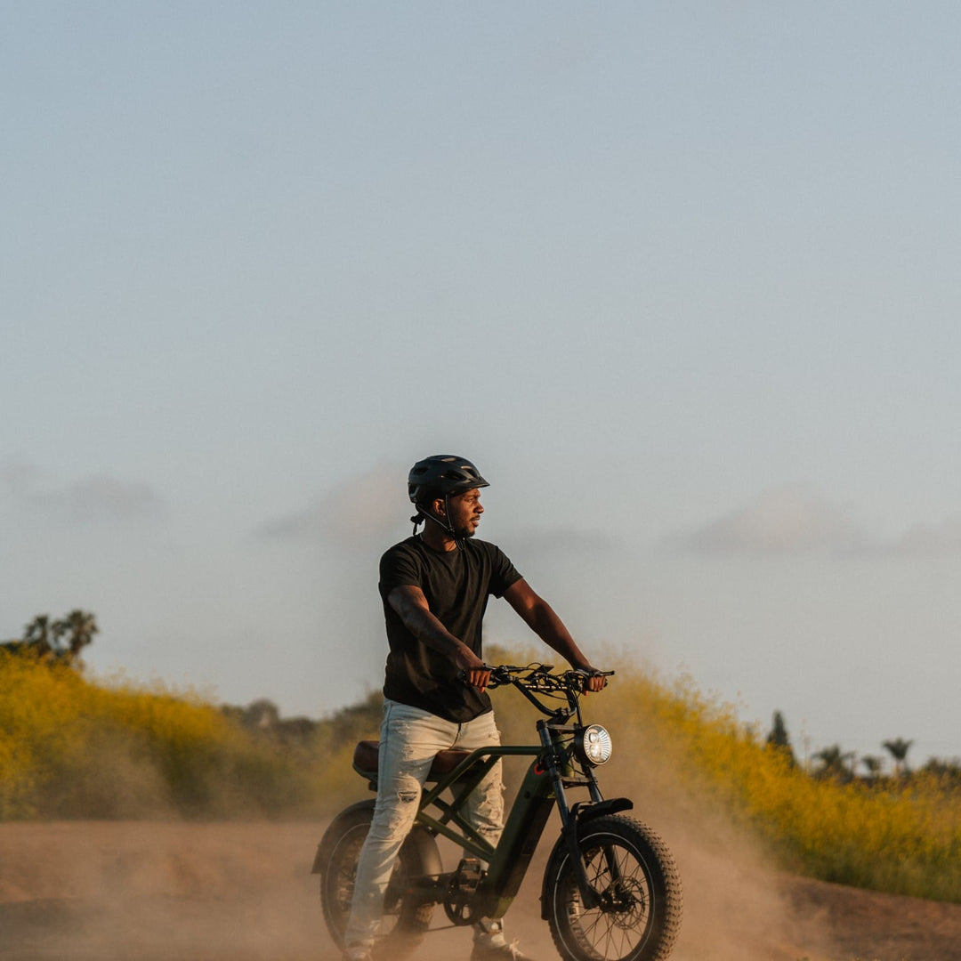 Man riding retrospec Valen Rev 20 fat tire electric bike on dusty trail with headlight, desert landscape.
