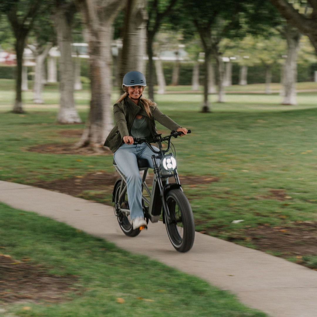 A smiling woman wearing a retrospec helmet rides the Valen Electric Bike toward the camera along a tree-lined park path, with the round LED headlight glowing and green lawns stretching behind her.