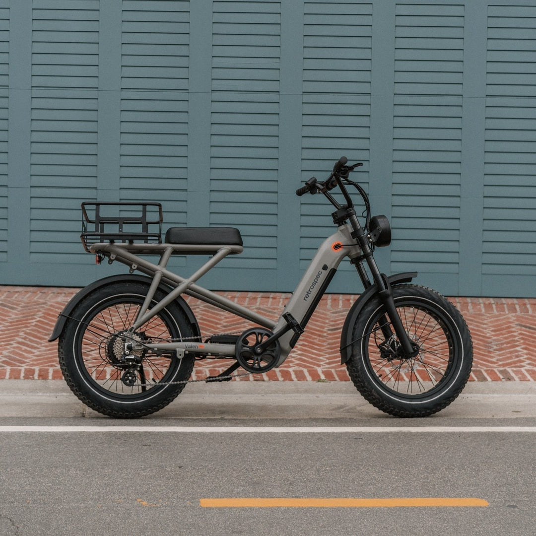 A sleek gray electric bike with a sturdy frame and wide tires parked beside a bright blue wall, featuring a brick pathway below.