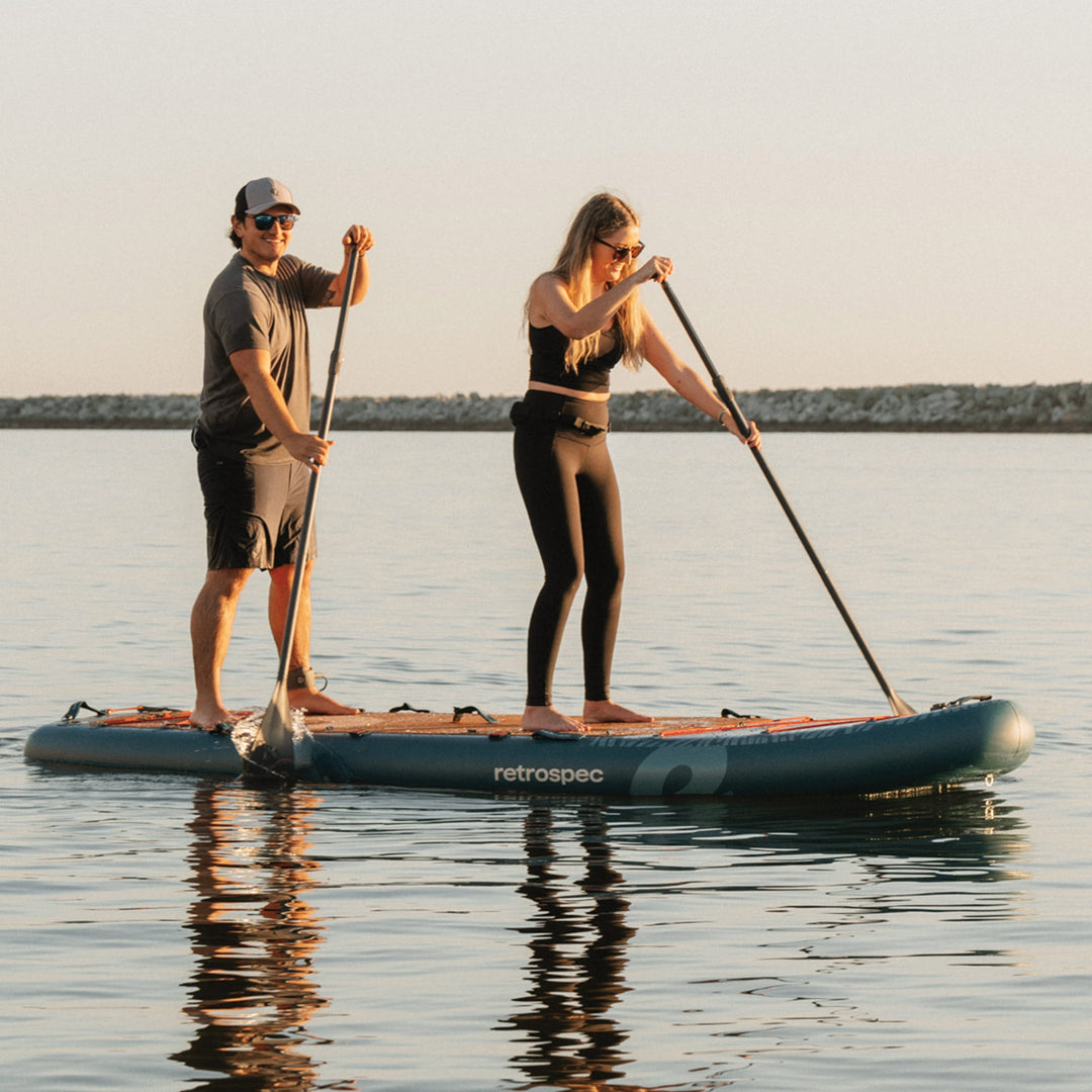 Man and woman paddling together on a dark teal retrospec tandem paddle board in calm, open water.