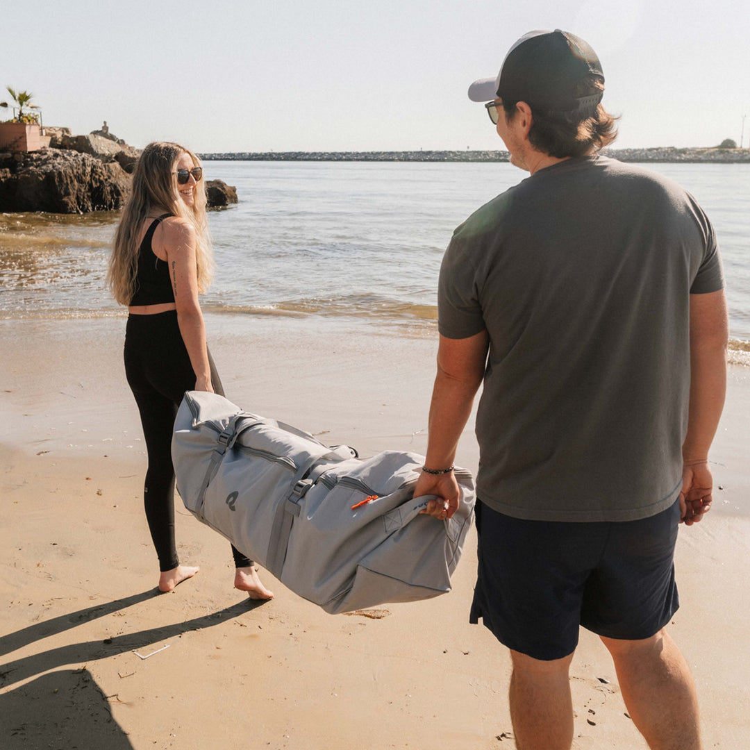 Two people smiling and carrying the retrospec Weekender Crew XL Paddle Board in its rolled travel bag along a sunny beach, with calm water and rocky breakwater in the background.