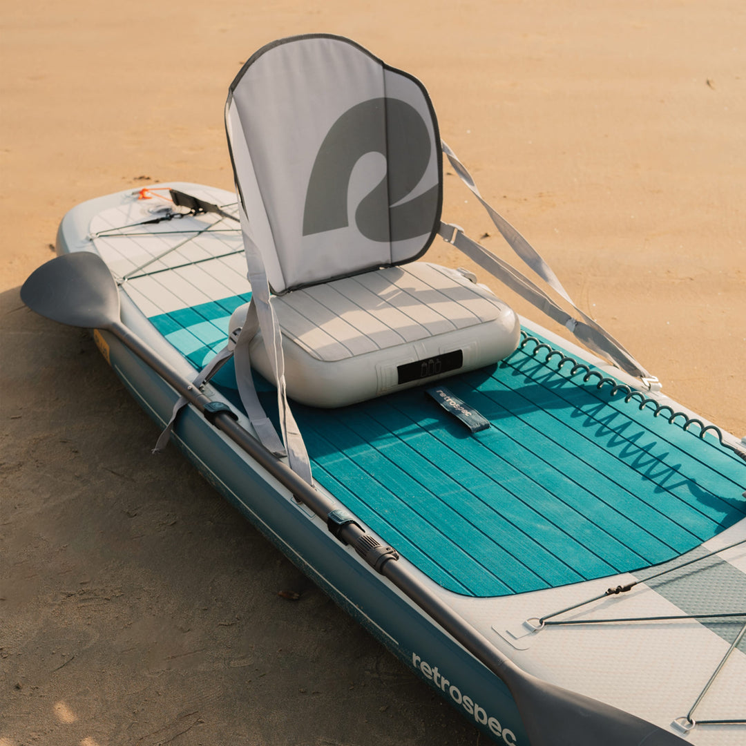 The Weekender Plus Inflatable Stand Up Paddle Board rests on the sand at the beach with the Kayak Seat and Paddle attached.