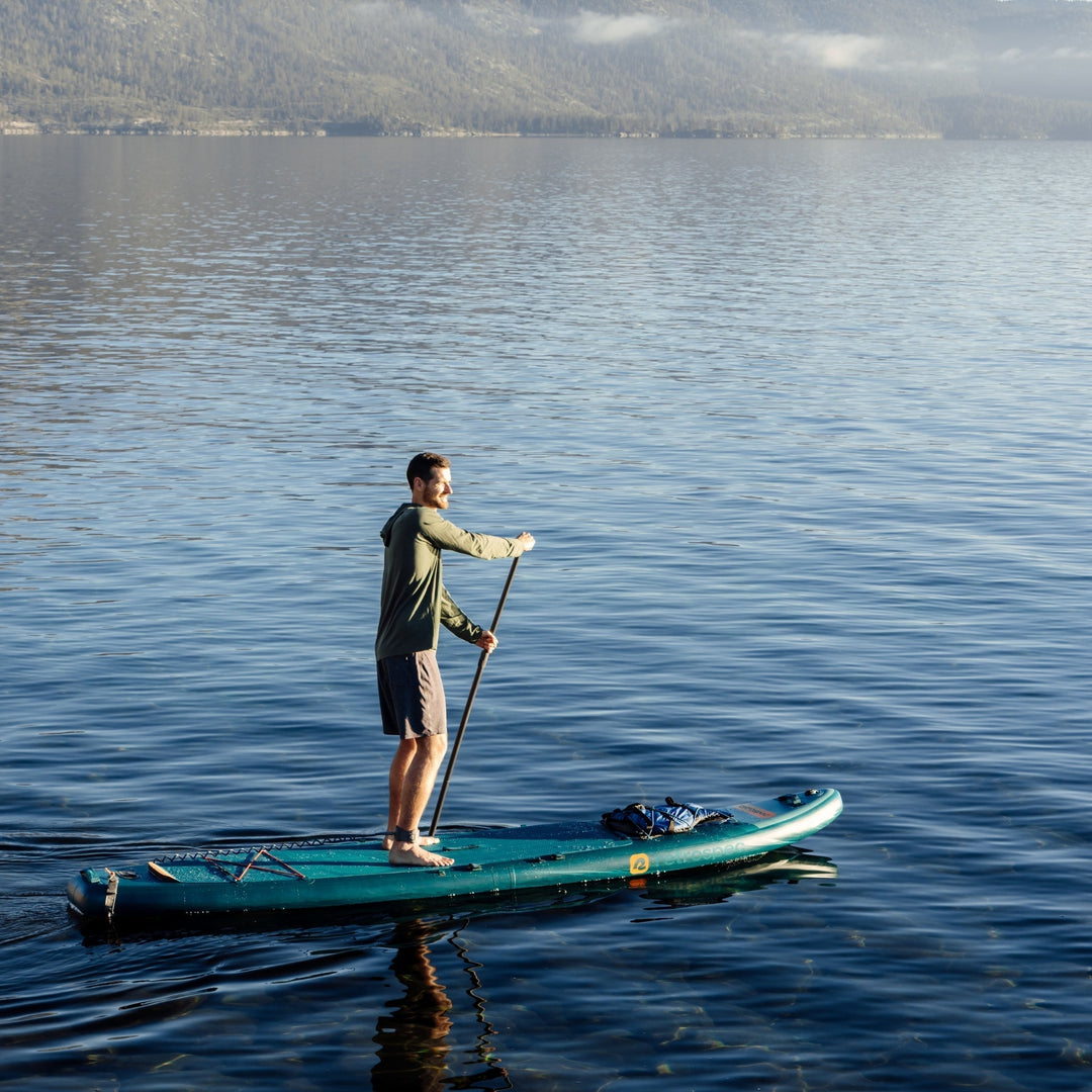 Person paddle boarding on teal weekender tour inflatable board on calm mountain lake with forested peaks in background