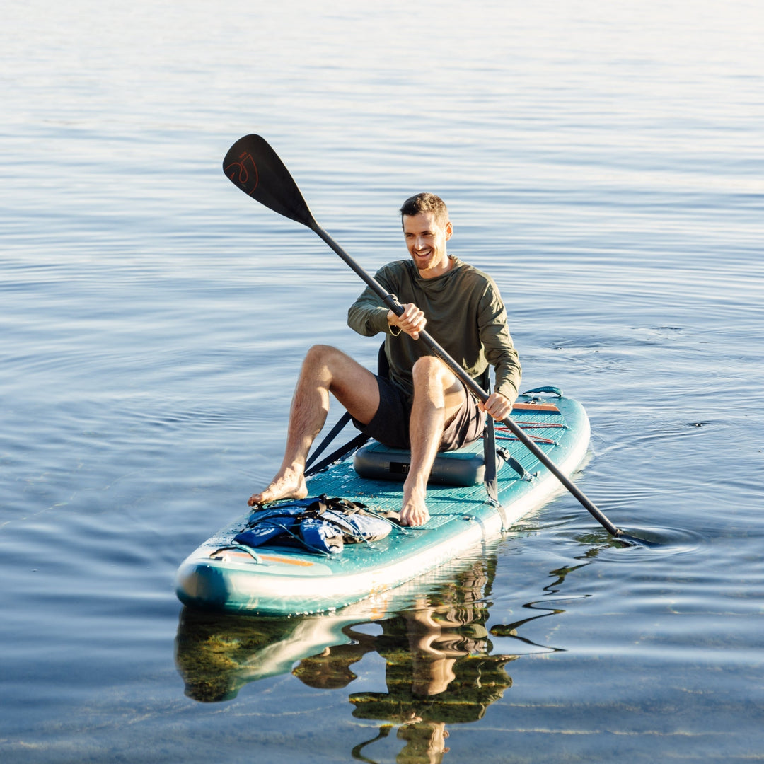 Smiling person paddling retrospec Weekender Tour XL stand up paddle board in teal on calm blue water with black paddle