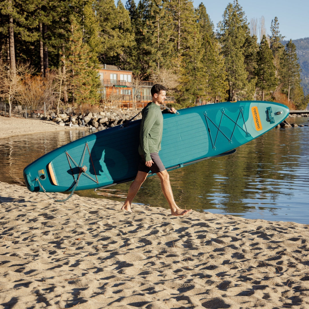 Person carrying retrospec Weekender Tour XL inflatable paddle board on sandy beach with forest and cabin in background