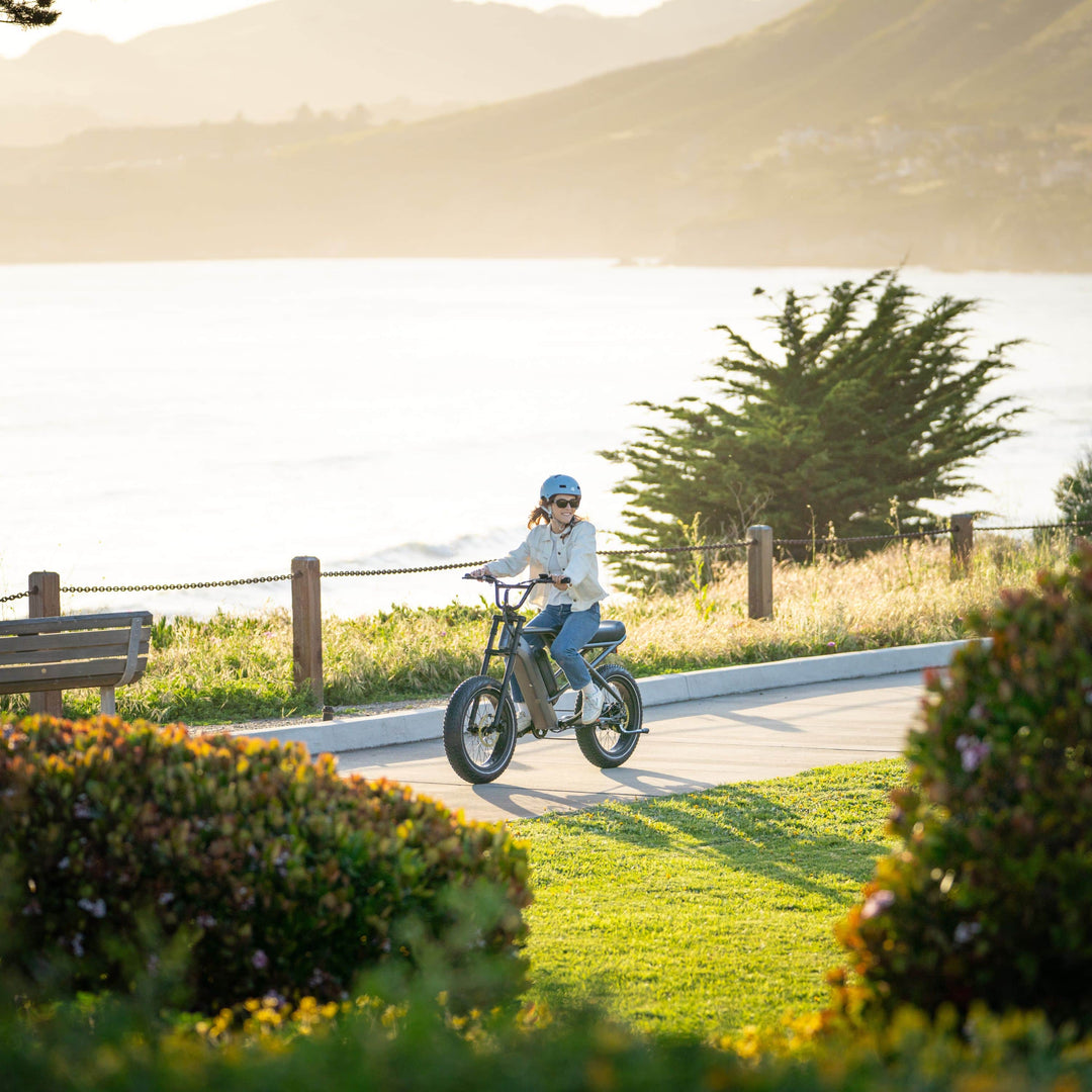 Woman riding a retrospec electric bike along a scenic coastal path with ocean views and wildflowers.