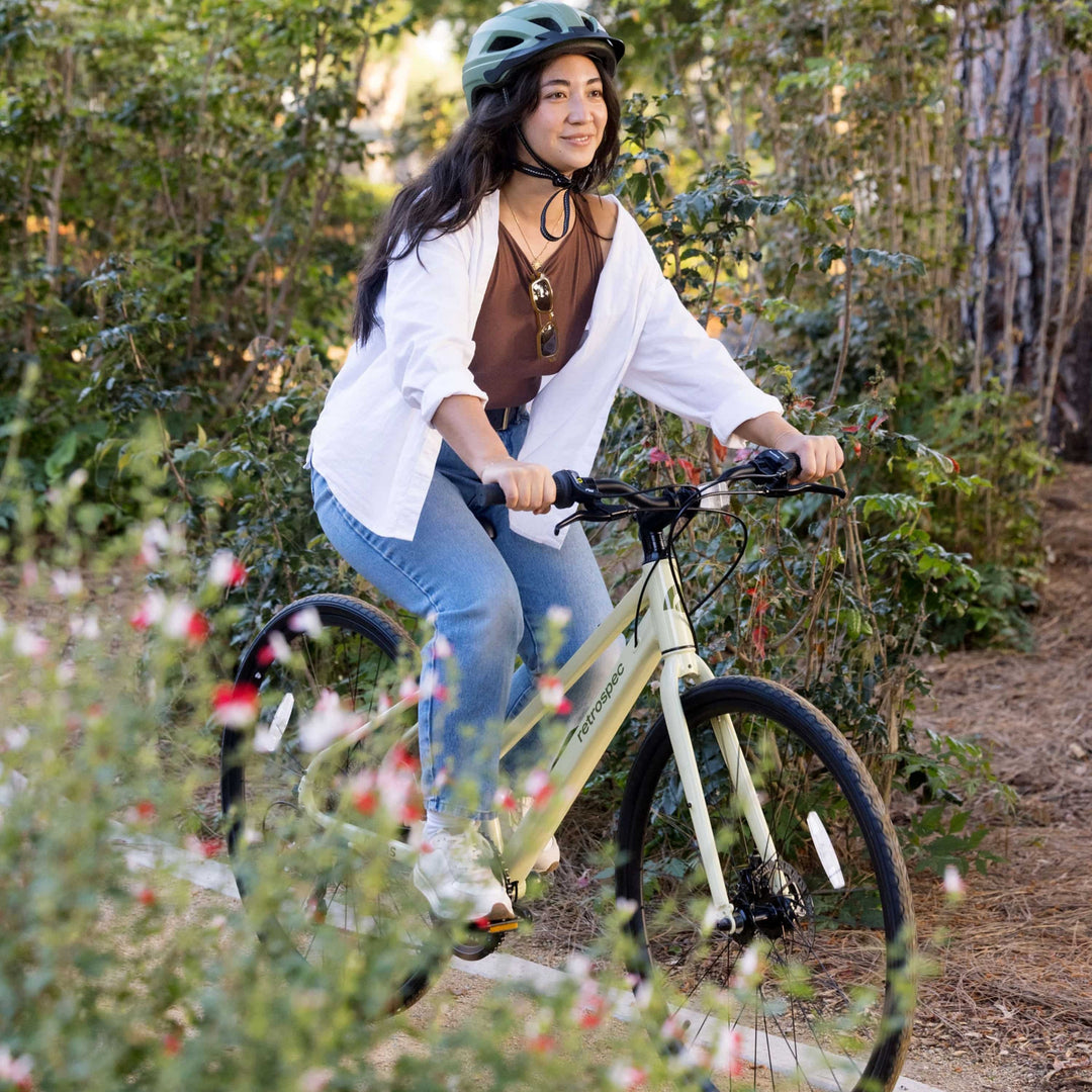A woman wearing a sage green Retrospec helmet rides a cream Retrospec Atlas hybrid bike along a lush garden trail surrounded by red and white wildflowers.