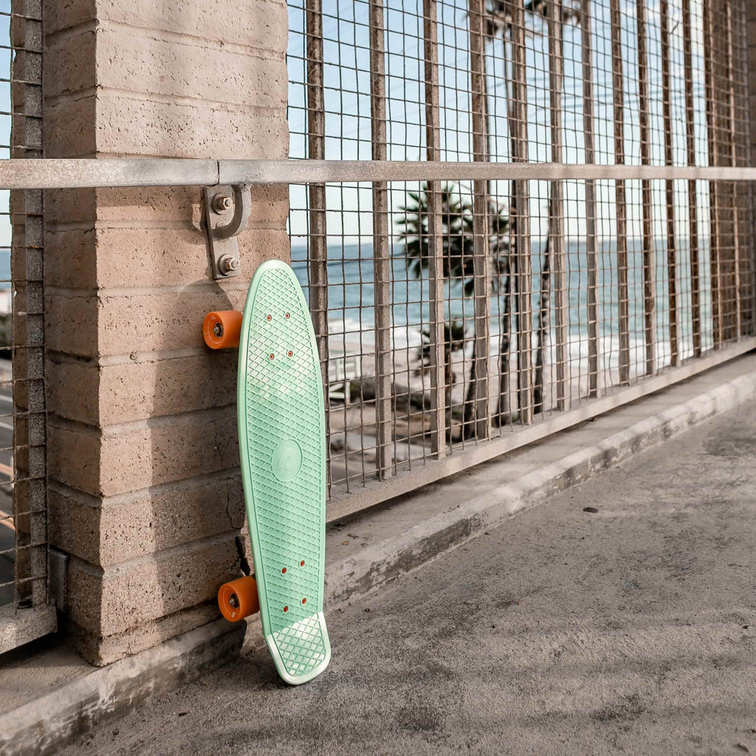 A mint green skateboard with orange wheels leans against a brick wall, overlooking the ocean behind a metal fence.