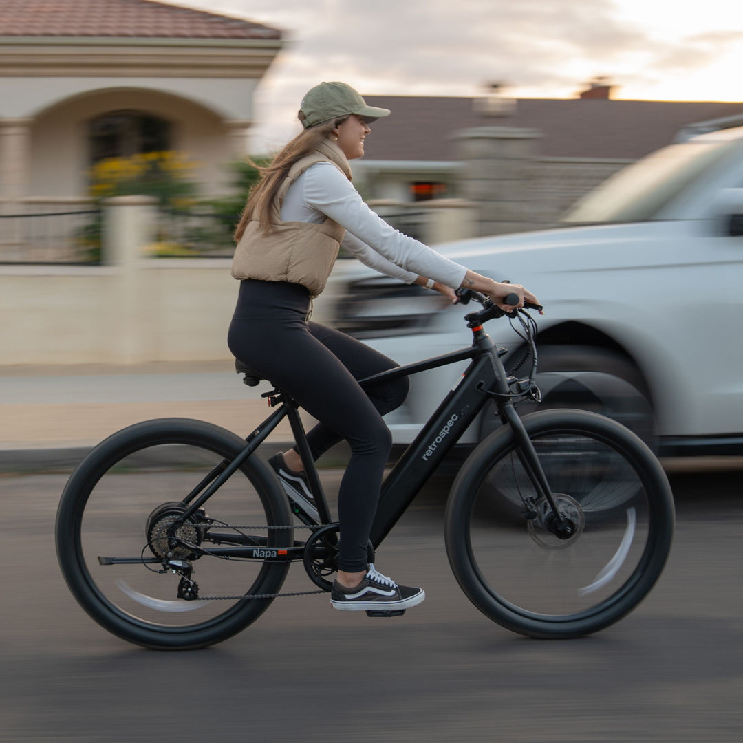 Woman smiling while riding a Retrospec Napa electric commuter bike down a city street at dusk with traffic in the background.