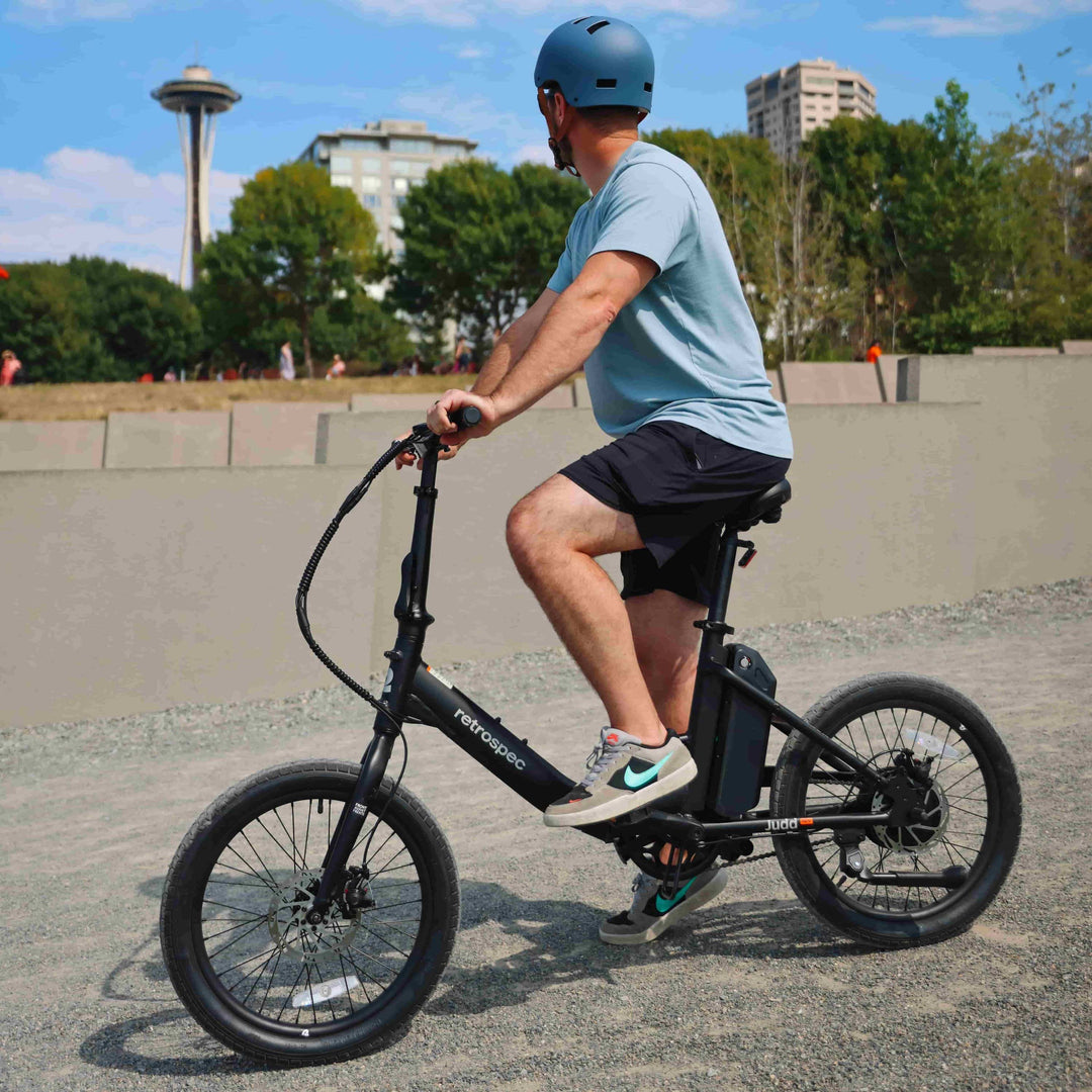 Man in blue helmet riding black retrospec Judra mini electric bike in urban park with Seattle Space Needle backdrop.