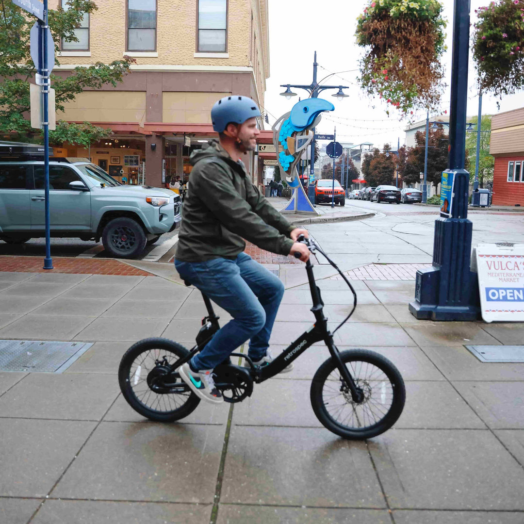 Man riding a retrospec Judd Rev Folding Electric Bike through a downtown street corner on a cloudy day.
