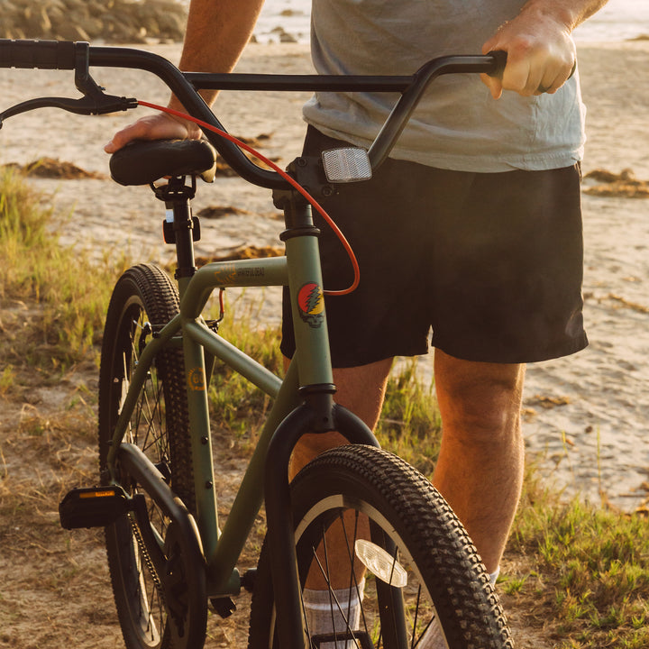 A person in a light blue shirt and black shorts holds a Grateful Dead Sully Klunker Bike by the handlebars, with a beach setting in the background.