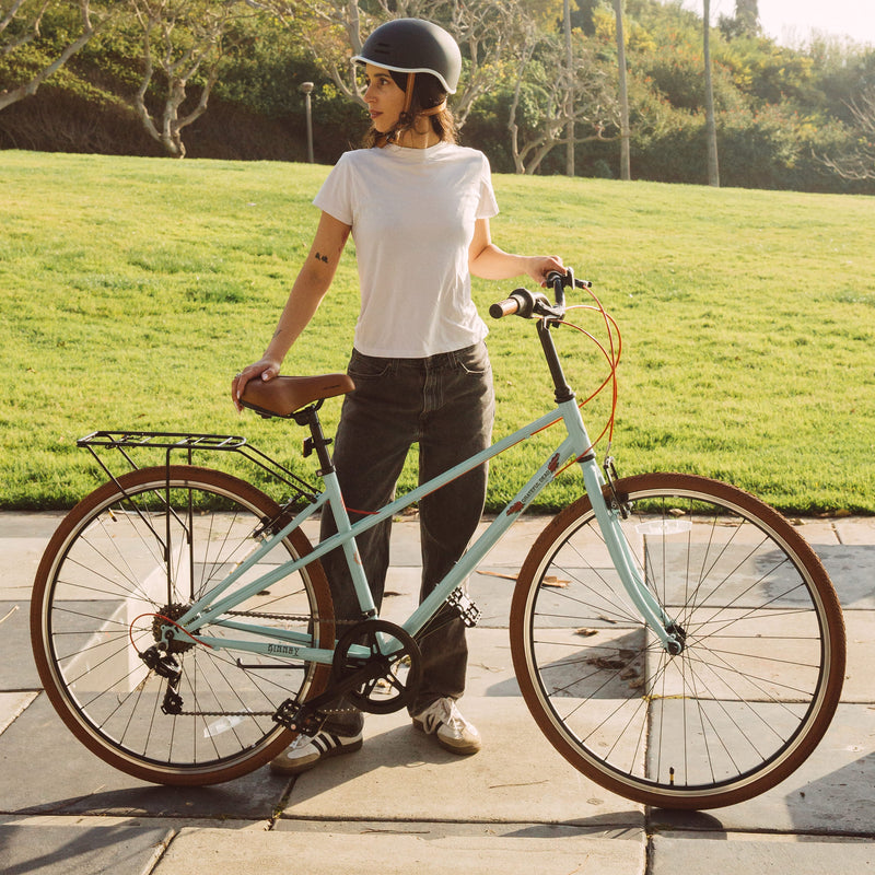 A person stands beside a seafoam Grateful Dead Kinney Mixte Bike with brown accents, wearing a white t-shirt and jeans, in a grassy outdoor setting.