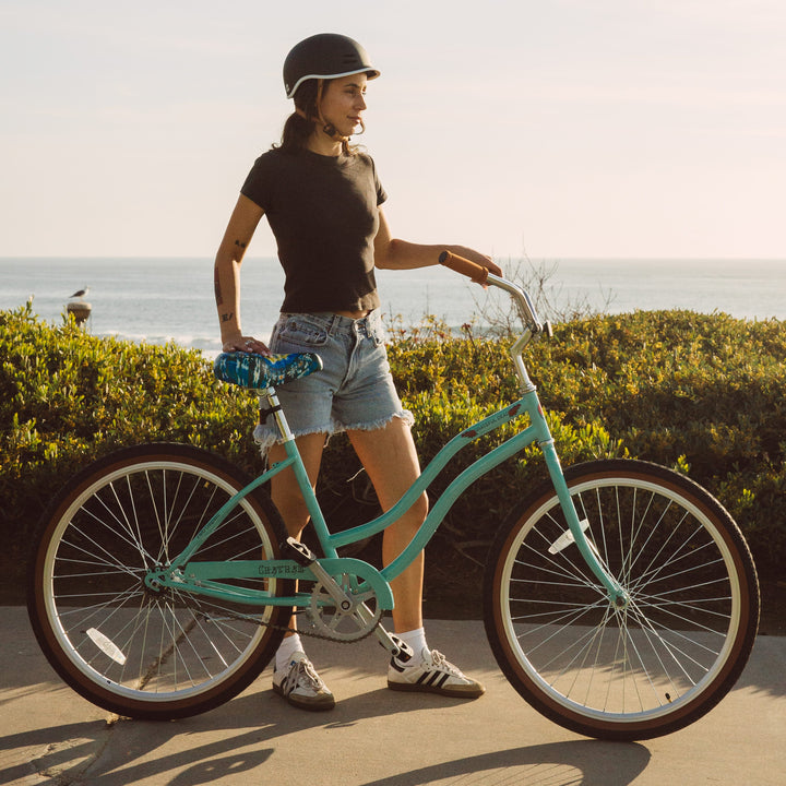 A person in a black t-shirt and denim shorts stands beside a turquoise Grateful Dead Chatham Single Speed Step Through Bike near the ocean, with greenery in the background.