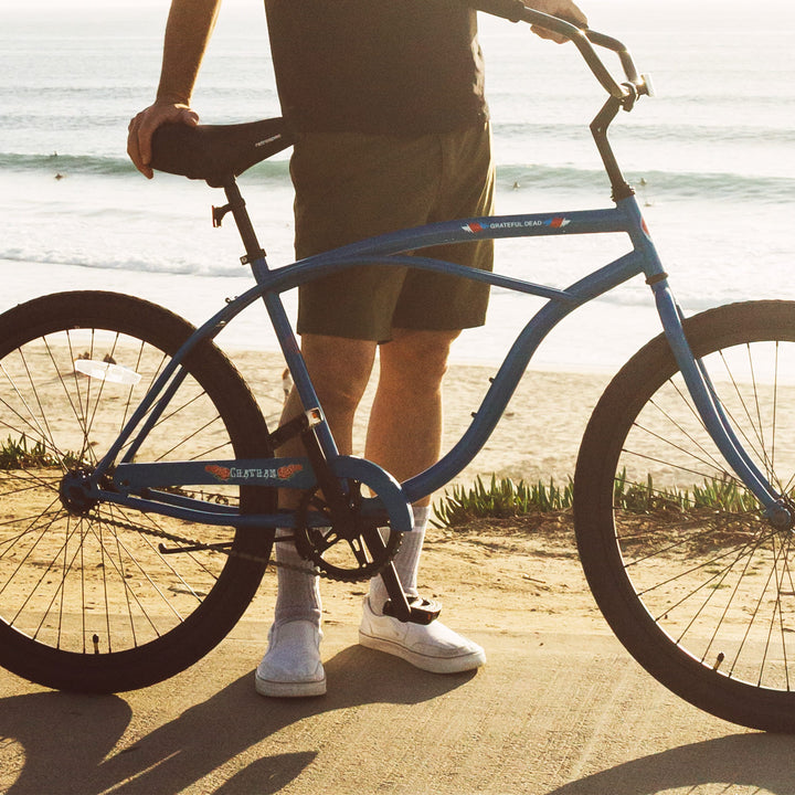 A person stands next to a blue Grateful Dead Chatham Single Speed Step Over Beach Cruiser Bike on a beachside path, with waves crashing in the background.