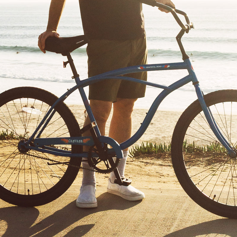 A person stands next to a blue Grateful Dead Chatham Single Speed Step Over Beach Cruiser Bike on a beachside path, with waves crashing in the background.