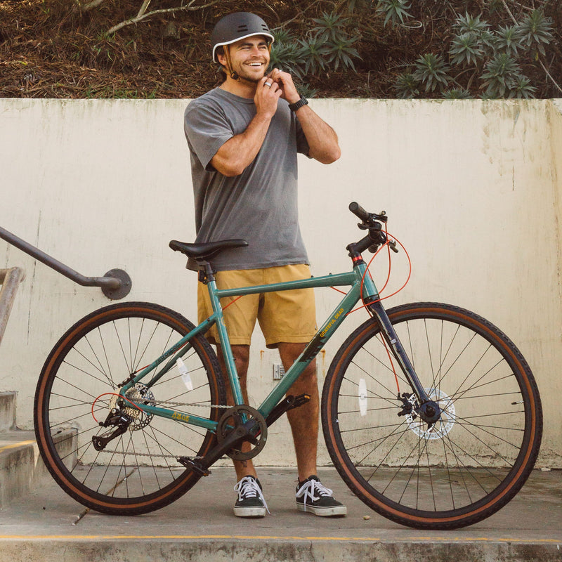 A person in a gray t-shirt and yellow shorts adjusts their helmet next to a green adventure bike on a stairway.