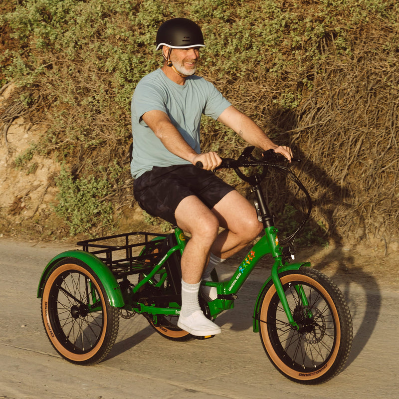 A person rides the bright green Grateful Dead Boca Rev Electric Tricycle on a dirt path, surrounded by lush greenery, wearing a helmet and casual clothes.