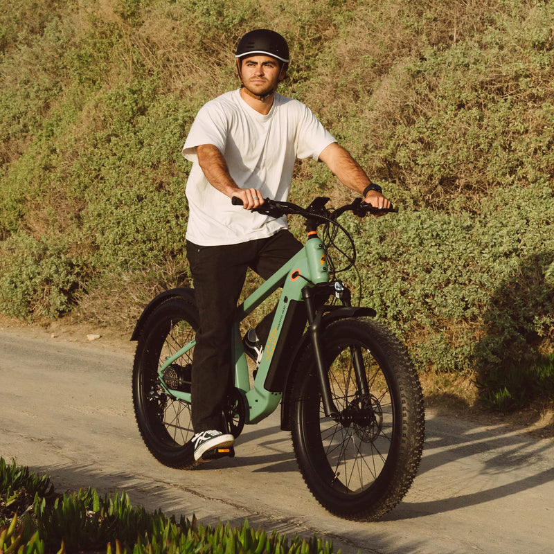 A person rides an electric bike on a gravel path surrounded by green foliage, wearing casual attire and a helmet.